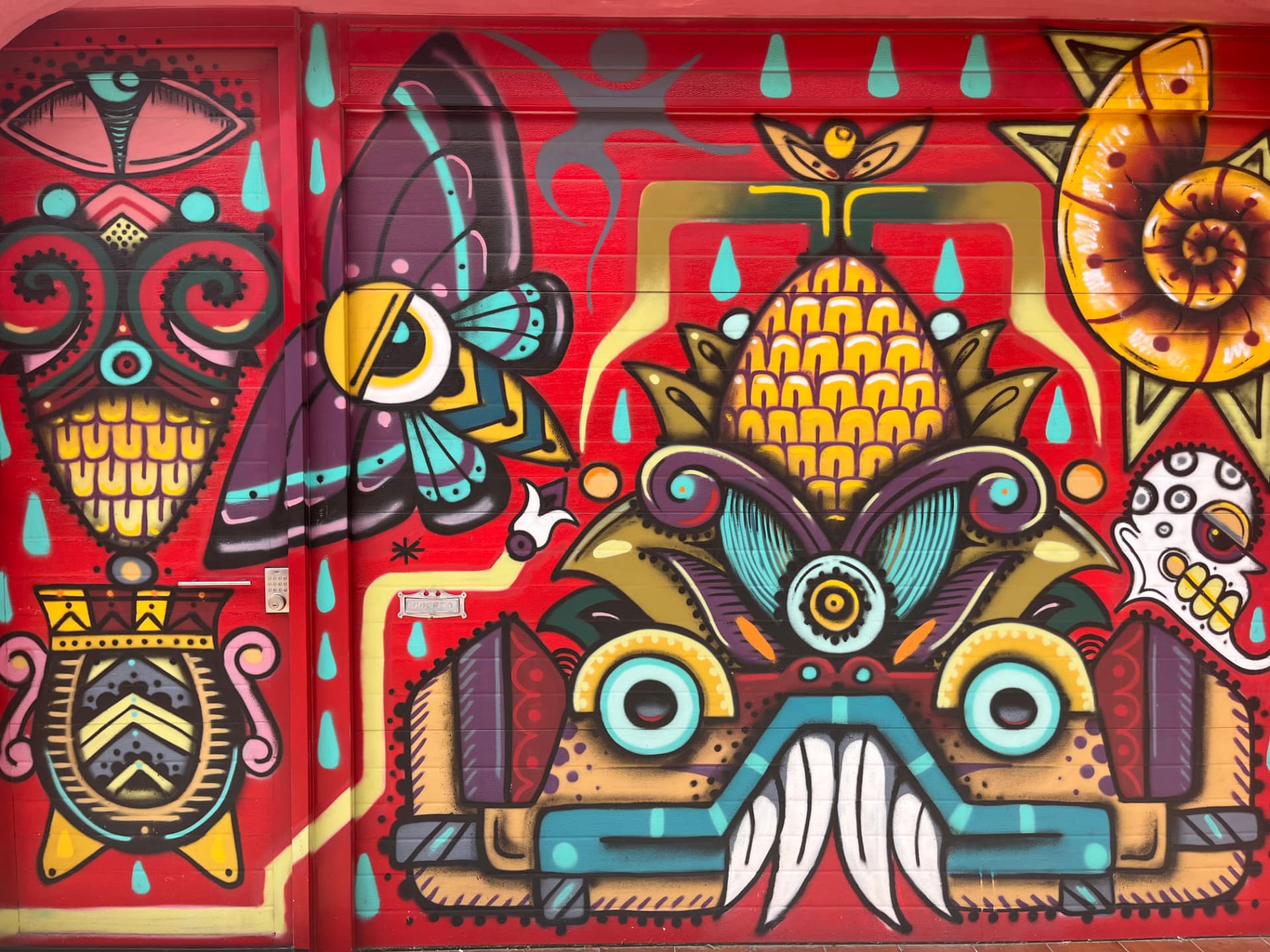 A vivid red street mural of a stylised mask and maize on a metal shutter, Mexico