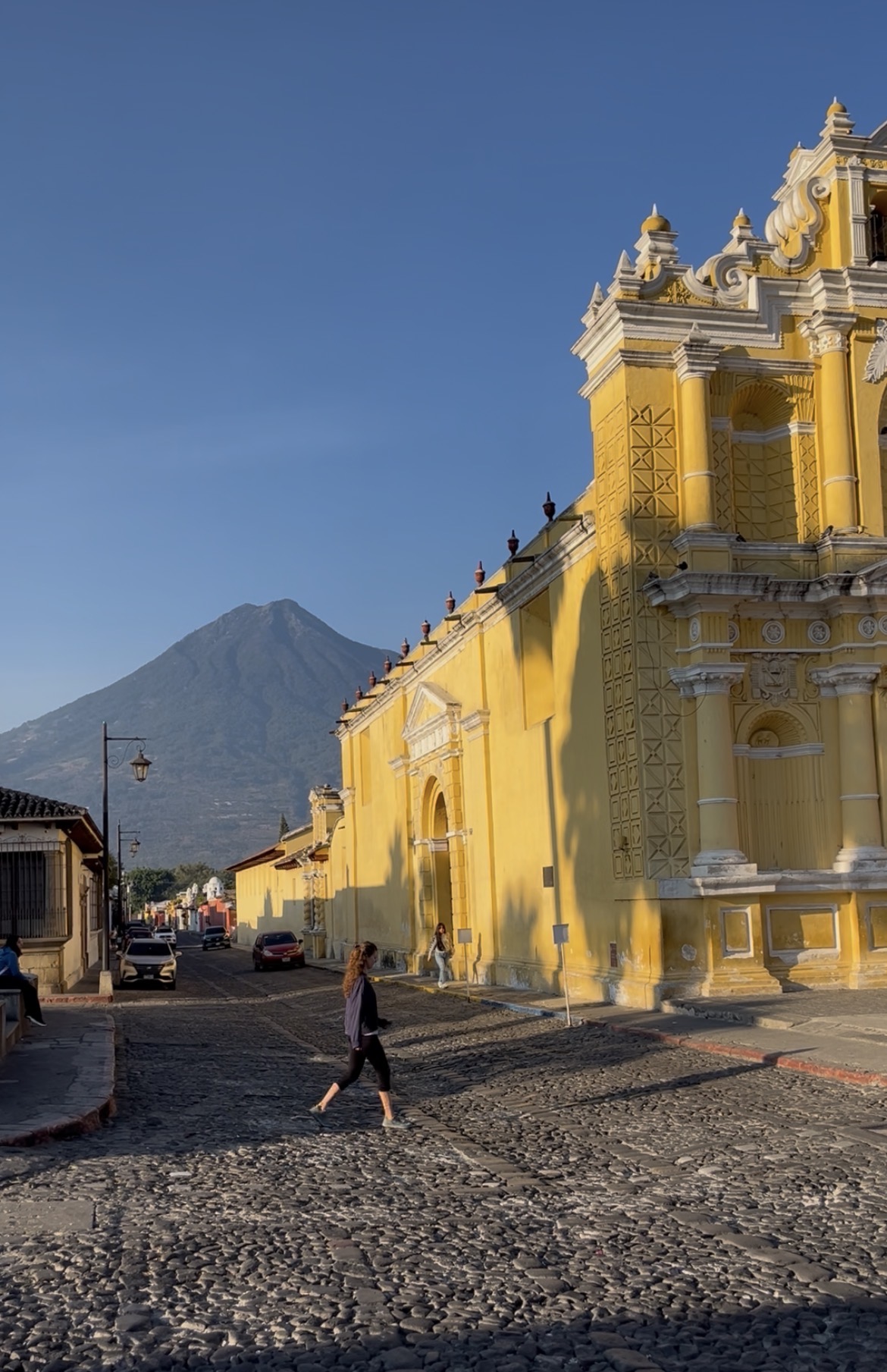 A person walking across a cobblestone street beside a golden church, a volcano beyond, Mexico