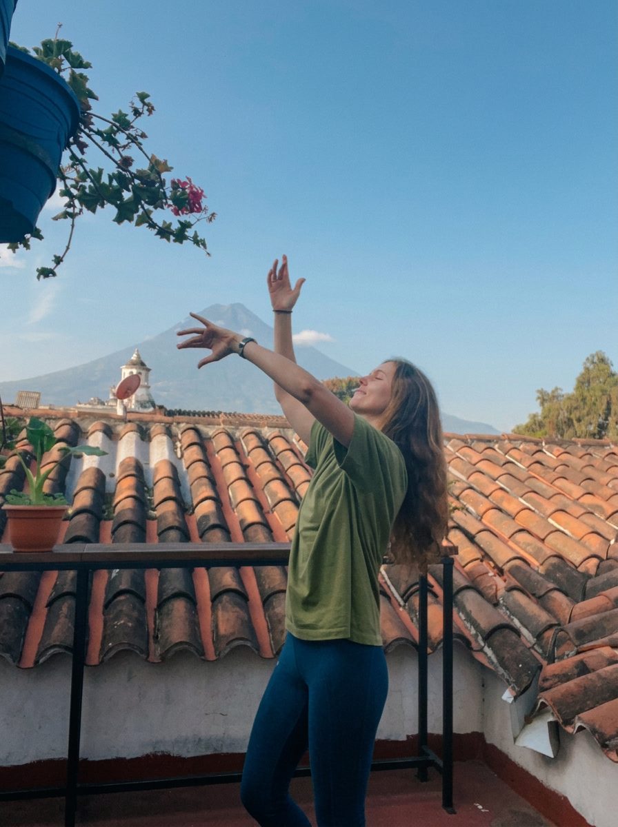 A woman on a rooftop reaching open arms toward the sky with a volcano in the distance